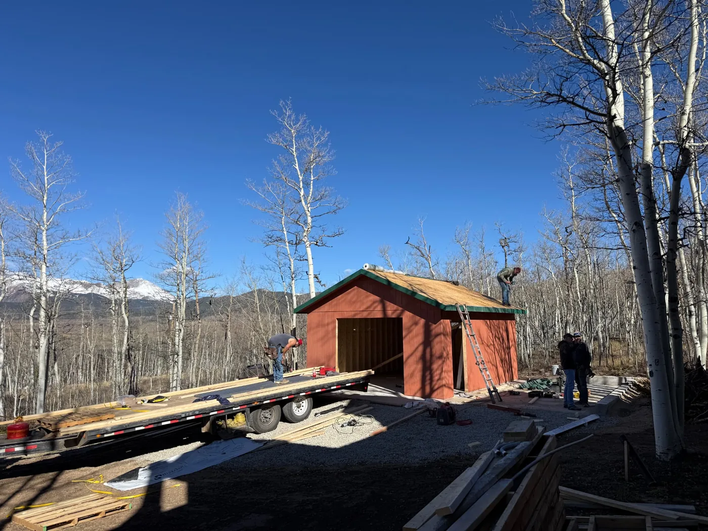 Crew members framing the roof of a terracotta-sided garage under construction, with roof decking installed, green fascia boards, a flatbed trailer and stacked lumber on site, and snow-capped peaks behind a stand of aspens