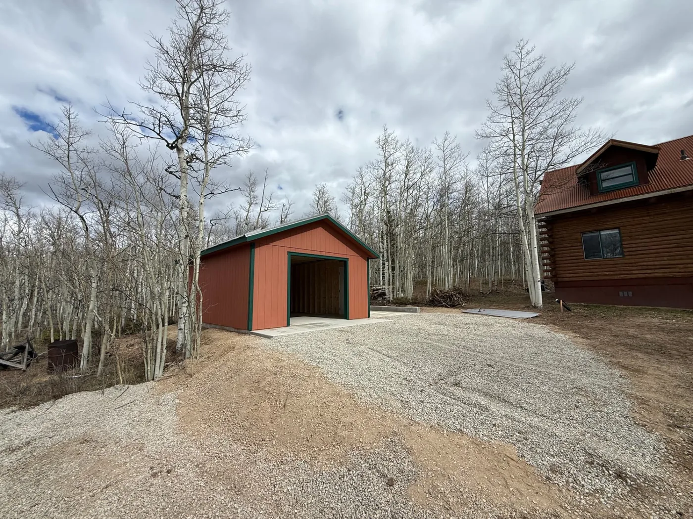 Front three-quarter view of a terracotta garage beside a log cabin, with light crushed road base spread across the graded approach and a dirt slope cut on the left