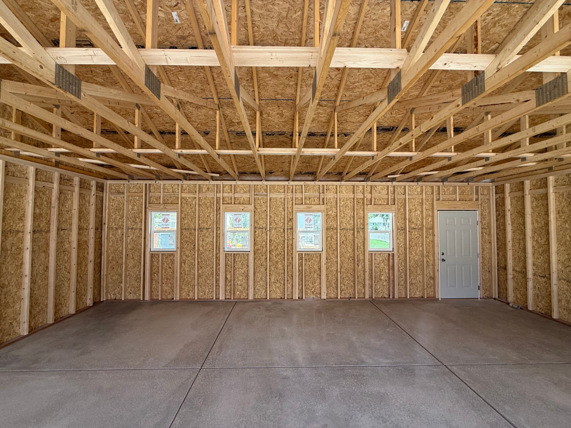 Unfinished garage interior with exposed wood framing, concrete floor, four rear windows and a pedestrian door