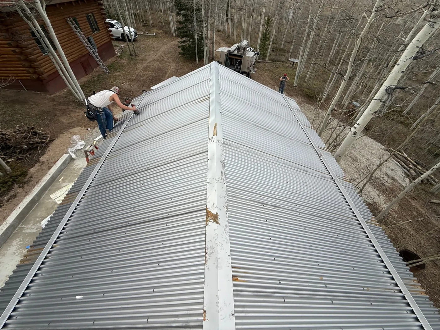 Overhead view looking down the ridge of a corrugated metal roof being installed, showing silver panels with rust-toned patina along the edges, rows of hex-head fasteners set into the ribs, and a metal snow break bar along the lower eave