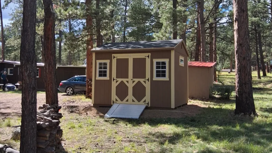 Tan 10x12 A Frame Shed with cream trim, black shingles, cream double doors, two windows and an aluminum ramp among pine trees