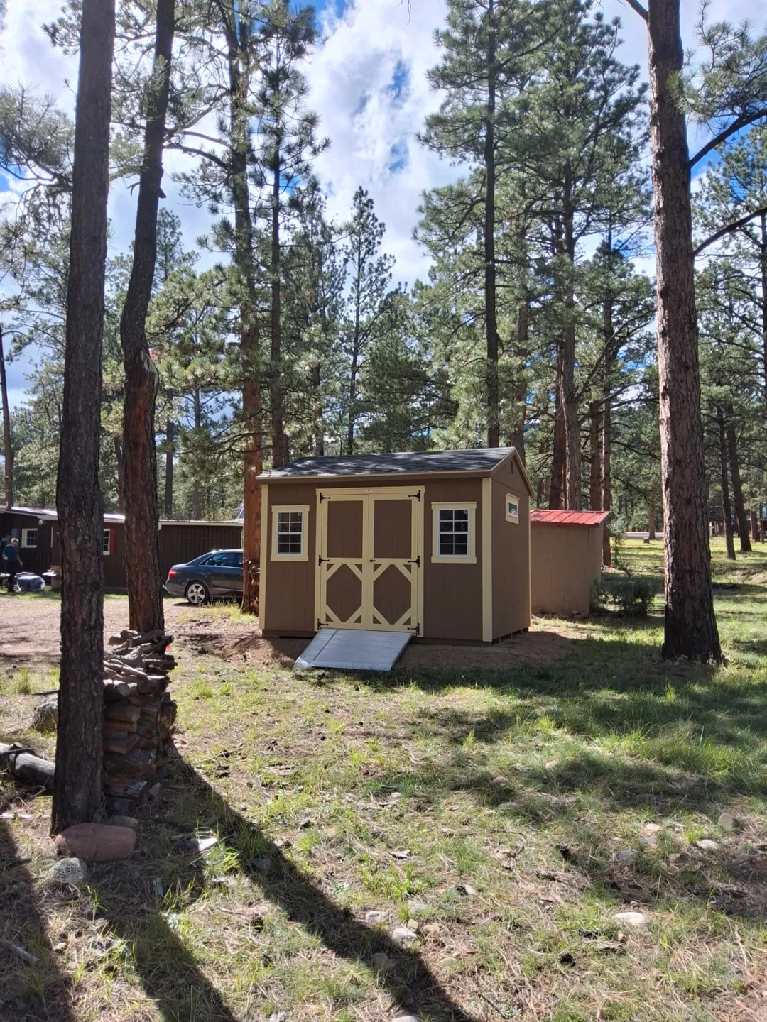 Tan 10x12 A Frame Shed with cream trim, black shingles, cream double doors, two windows and an aluminum ramp among pine trees