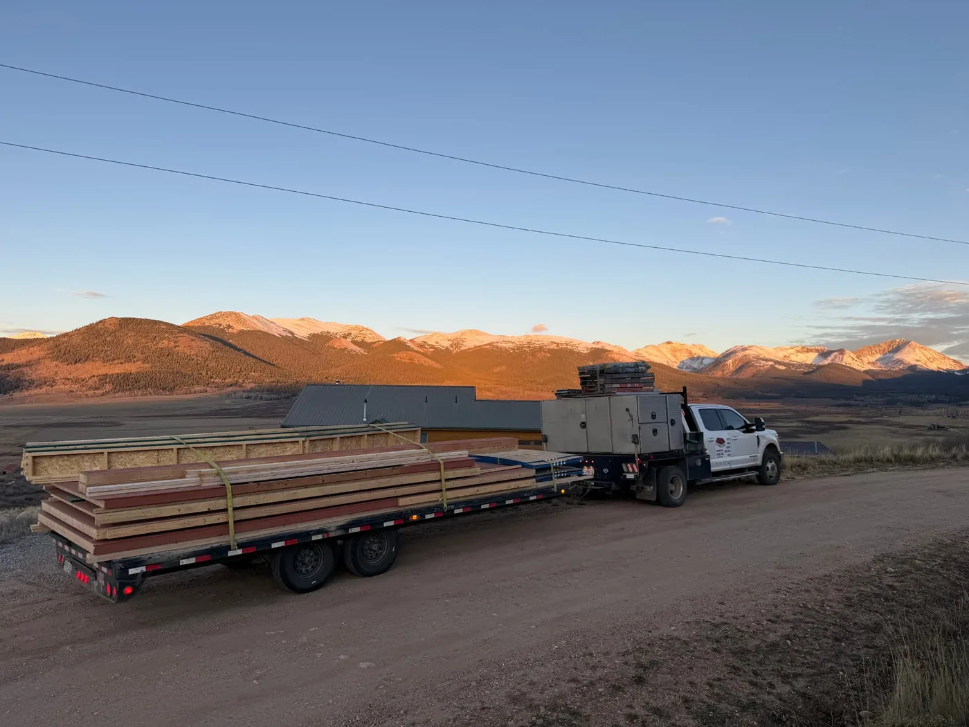 White flatbed pickup hauling banded prefabricated wall panels and framing lumber on a tandem-axle trailer along an unpaved county road, with a wide valley and snow-dusted peaks in golden light behind