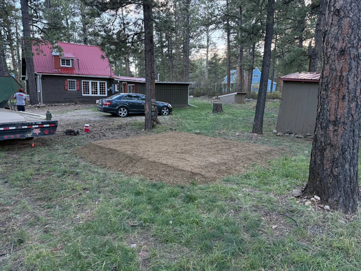 Freshly prepared rectangular gravel road base pad on a grassy, pine-shaded lot, with a trailer and work crew in the background
