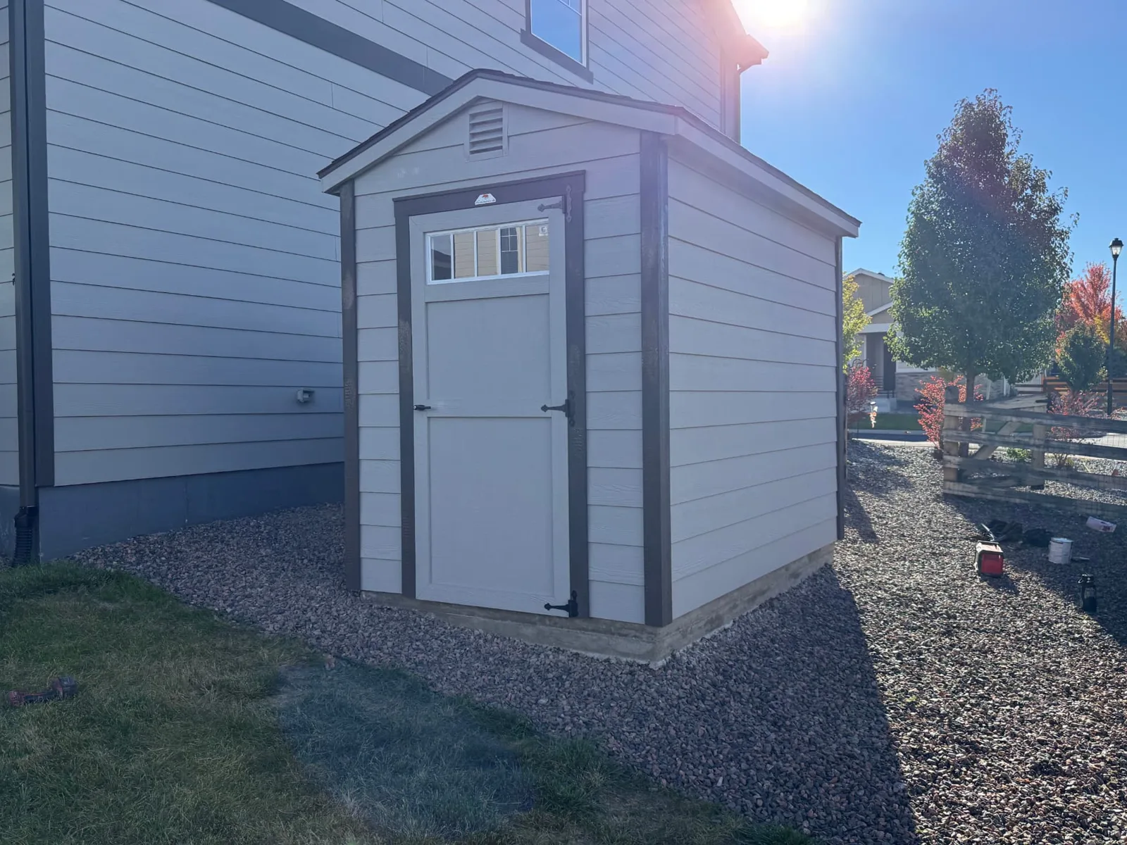 Front and side of the gray shed showing lap siding, dark trim and a wood door with a transom window