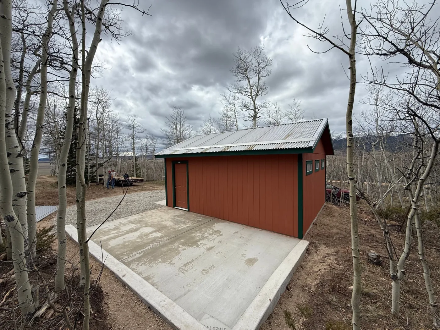 Rear and side view of a completed terracotta garage with dark green trim, a painted steel entry door, three transom windows, and a broom-finished concrete slab bordered by a raised curb, with a gravel turnaround and mountains visible through the trees